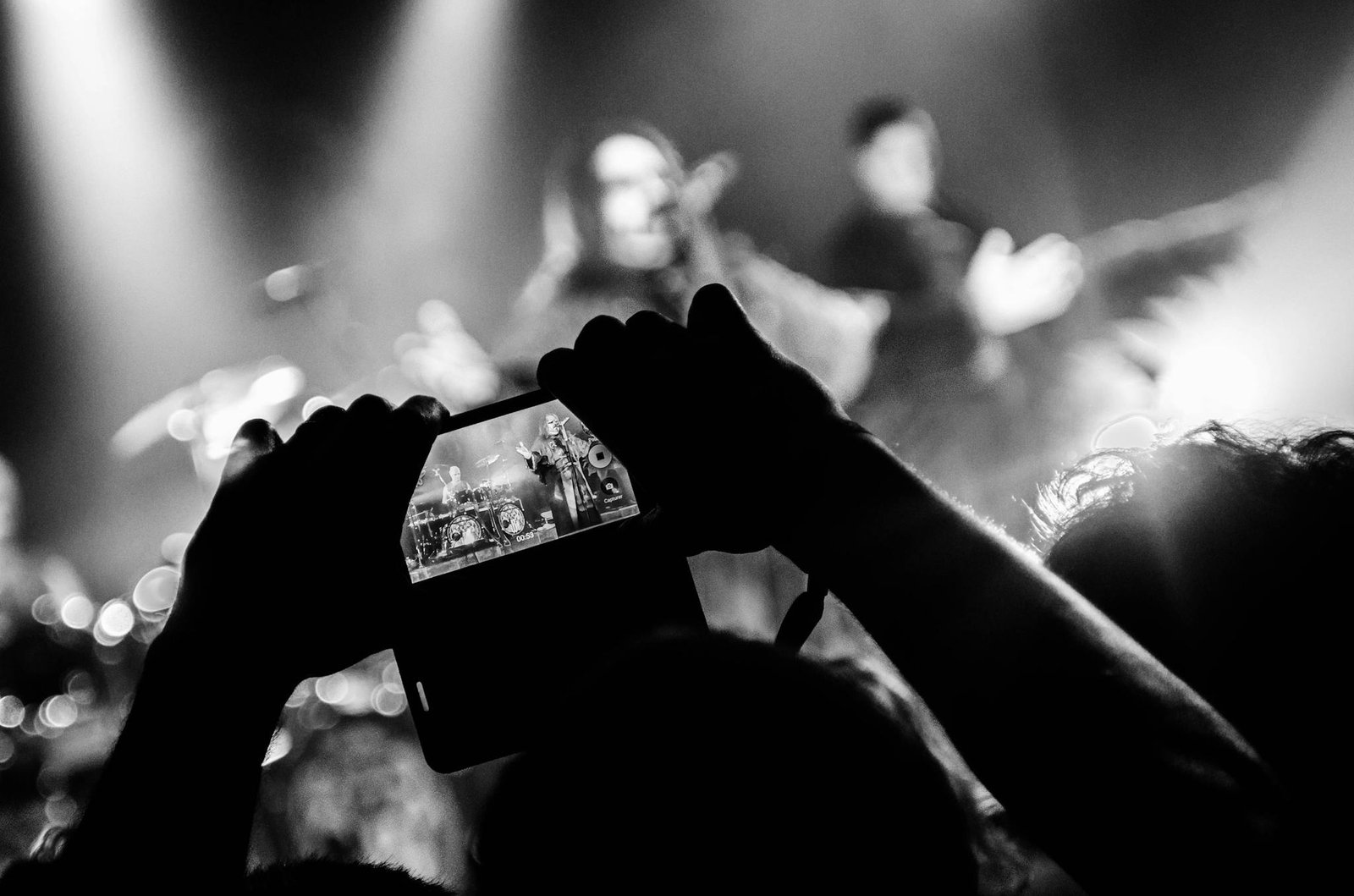 Black and white photo of a concert with a focus on filming the band using a smartphone.